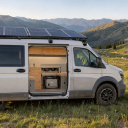 Bus aménagé autonome stationné dans une prairie naturelle, avec des panneaux solaires sur le toit et un intérieur en bois clair laissant voir une batterie auxiliaire.